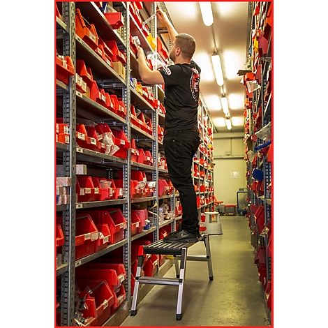 Ein Mann steht auf einer Trittleiter und greift in ein Regal voller roter Behälter. Er trägt ein schwarzes T-Shirt und eine schwarze Hose.